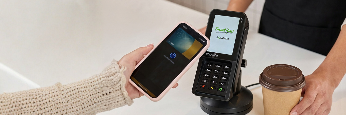 equinox-luxe-6700x-mobile-payment-apple-pay-qsr Customer using Apple Pay on a smartphone to tap and pay at an Equinox Luxe 6700x payment terminal at a quick-service coffee shop counter.